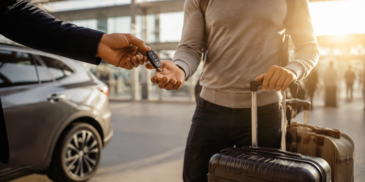 Close-up of a car key being handed between two people near a parked car with luggage